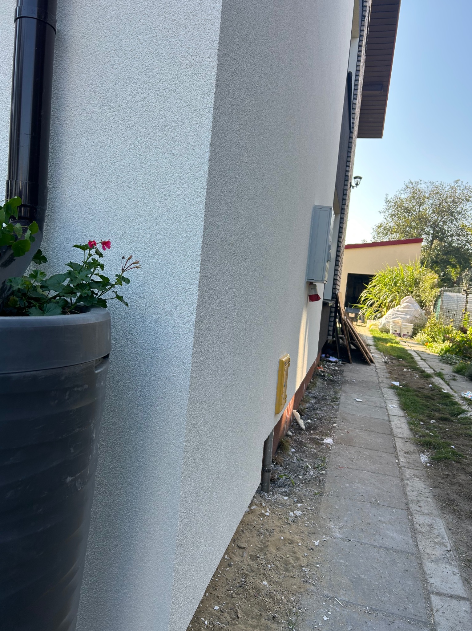 Biała elewacja domu po renowacji, widok z boku z donicą z kwiatami i fragmentem chodnika. Widoczne detale faktury tynku i elementy instalacji zewnętrznej.
