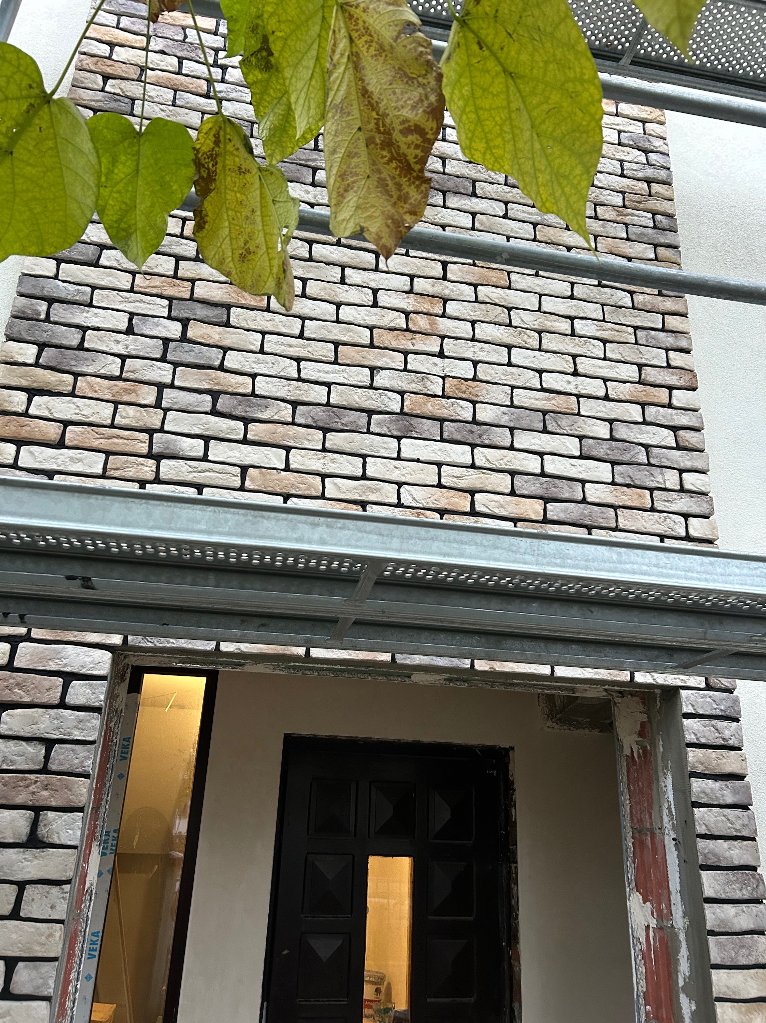 Elewacja budynku z dekoracyjną okładziną z cegły, widoczne rusztowanie i fragment roślinności. Detal wykończenia w trakcie prac.