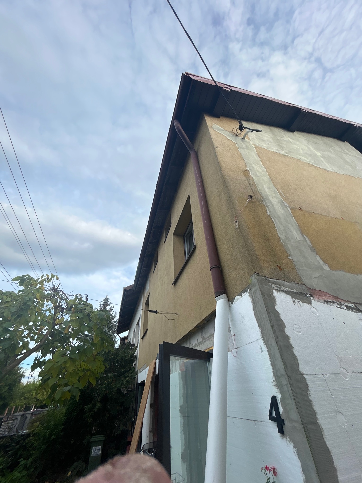 Dom w trakcie ocieplania: styropian na ścianie, numer '4', częściowo otynkowana elewacja, brązowa rynna. Widok z dołu, pochmurne niebo w tle.