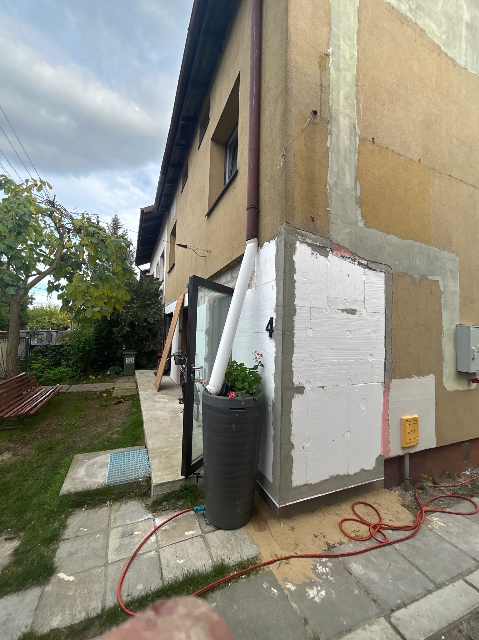 Ocieplenie fragmentu ściany budynku styropianem, widoczne ślady po szpachlowaniu, rura spustowa i donica z kwiatami. Prace w toku, widok z zewnątrz.