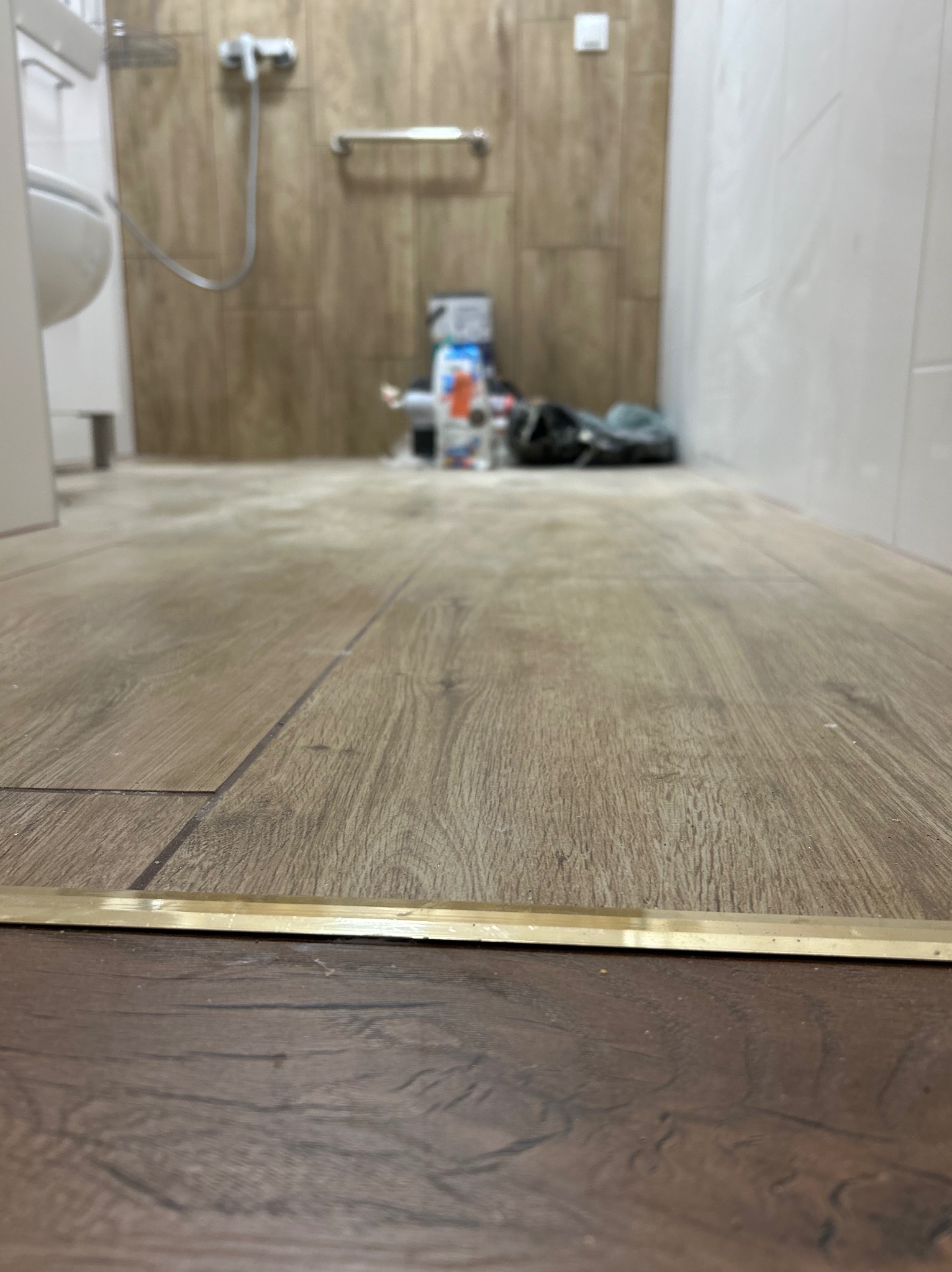 Ukończona podłoga w łazience z panelami imitującymi drewno, widoczna listwa progowa. W tle fragment toalety i ściany wyłożonej płytkami.