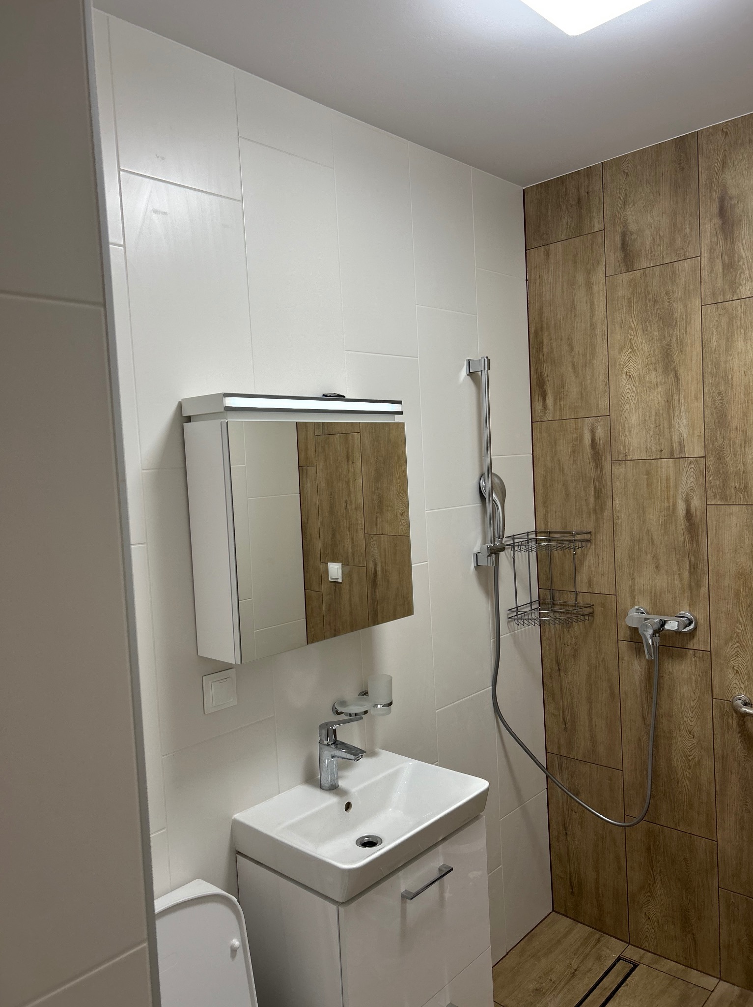 Nowoczesna łazienka z białymi płytkami i drewnopodobnymi panelami. Umywalka z szafką, lustro z oświetleniem, kabina prysznicowa z deszczownicą.