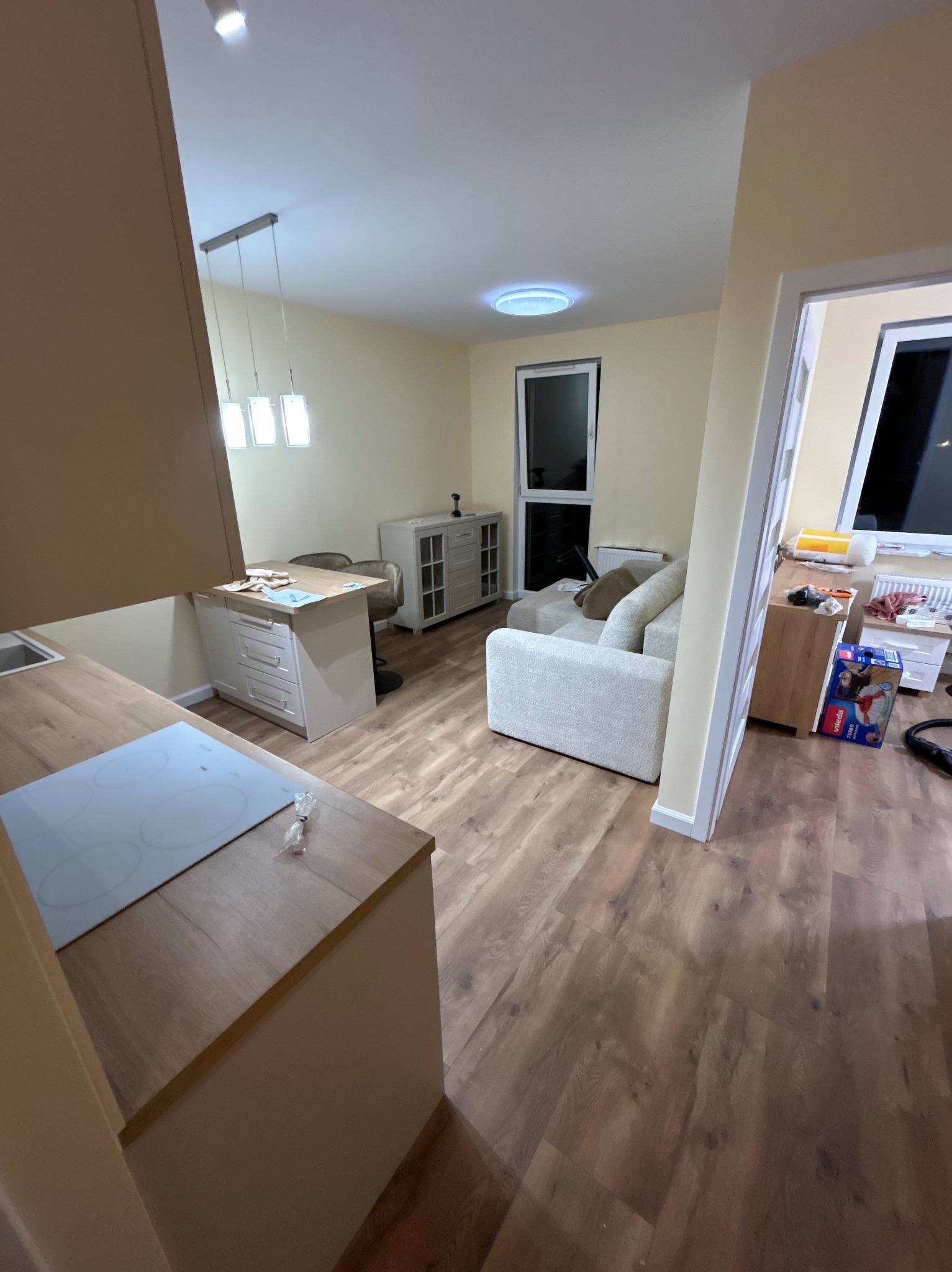 Jasne, przestronne wnętrze po remoncie: drewniana podłoga, aneks kuchenny z płytą indukcyjną, salon z sofą i oknem balkonowym. Nowoczesne oświetlenie sufitowe.