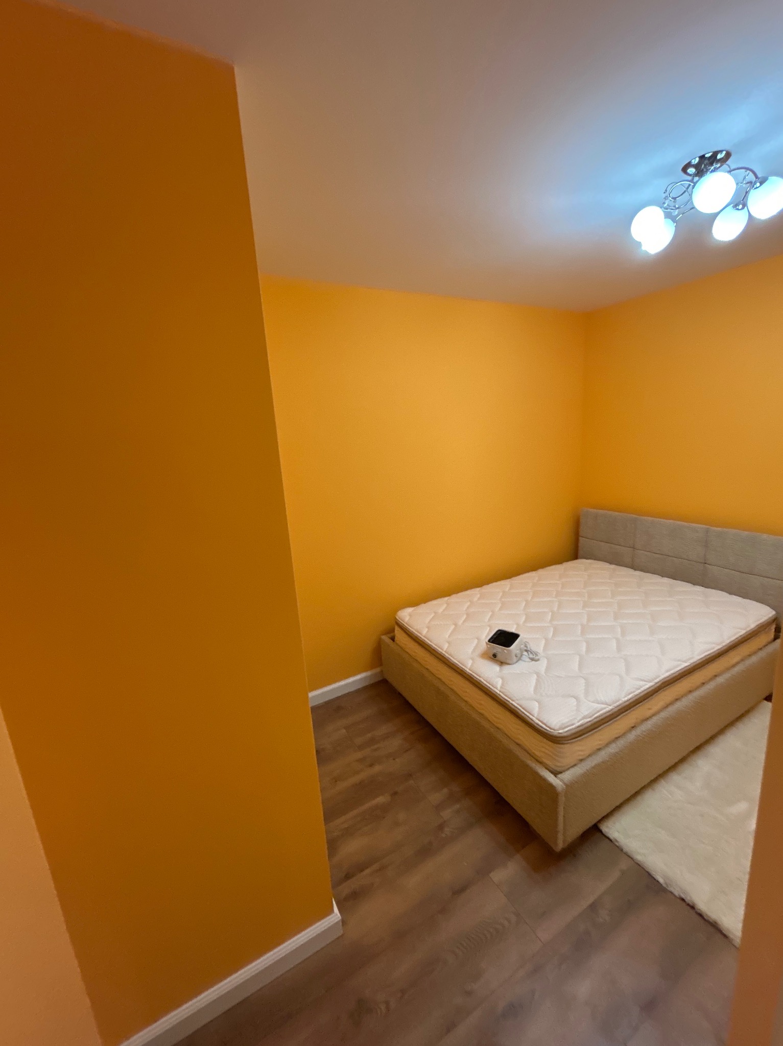 Pokój z łóżkiem w odcieniach pomarańczy i beżu. Ściany pomalowane na ciepły odcień pomarańczowego, drewniana podłoga i beżowe łóżko z białym materacem. Nowoczesna lampa sufitowa.