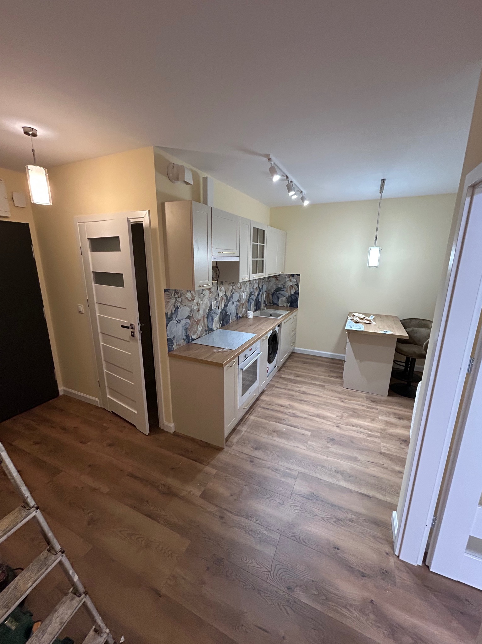 Jasna kuchnia po remoncie z drewnianą podłogą, wzorzystym panelem nad blatem i białymi szafkami. Widoczny stół z krzesłem i drabina malarska.