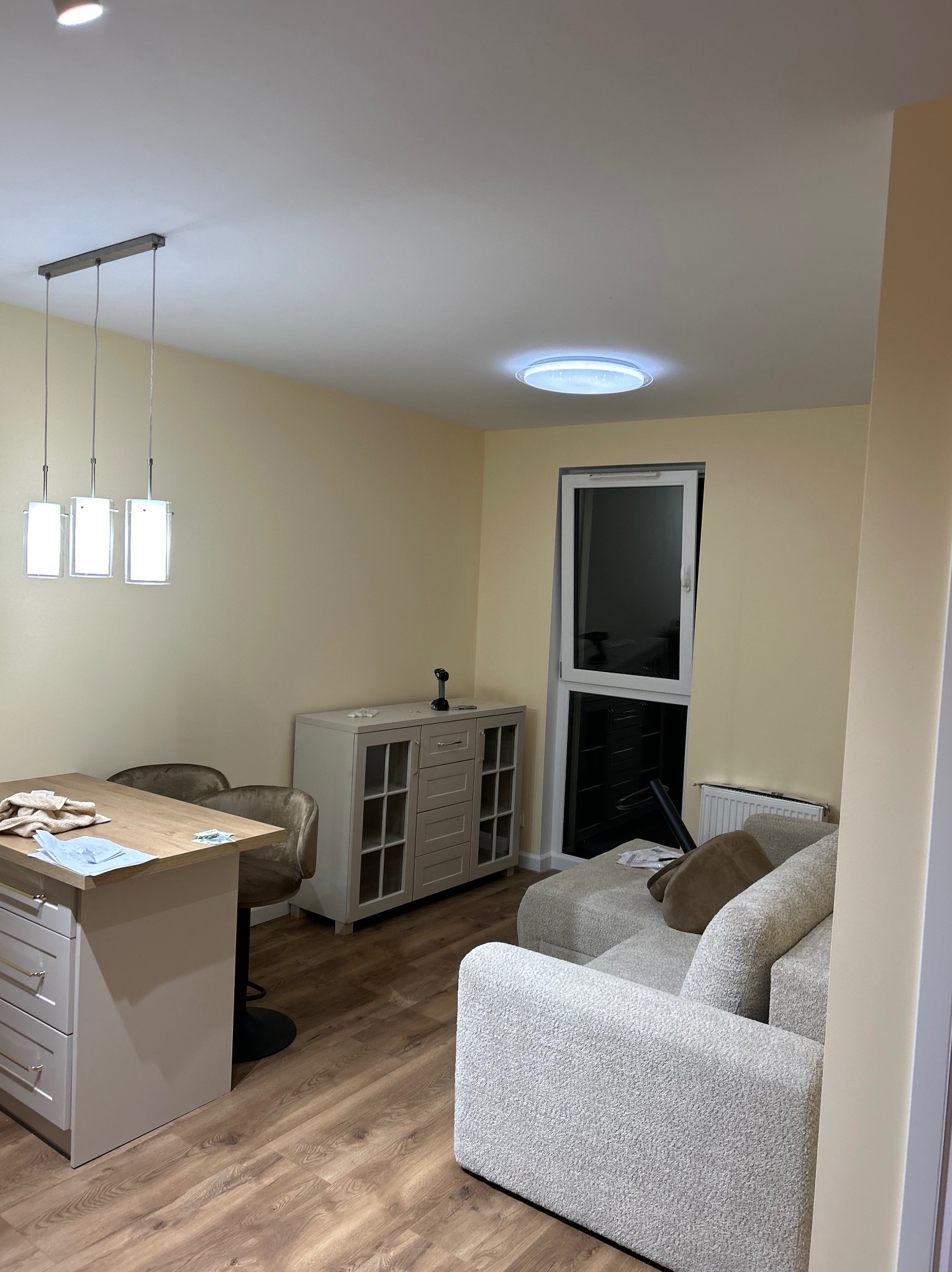 Jasne wnętrze po odświeżeniu: kremowe ściany, drewniana podłoga, nowoczesne oświetlenie nad wyspą kuchenną i wygodna sofa w salonie. Widoczny fragment okna balkonowego.