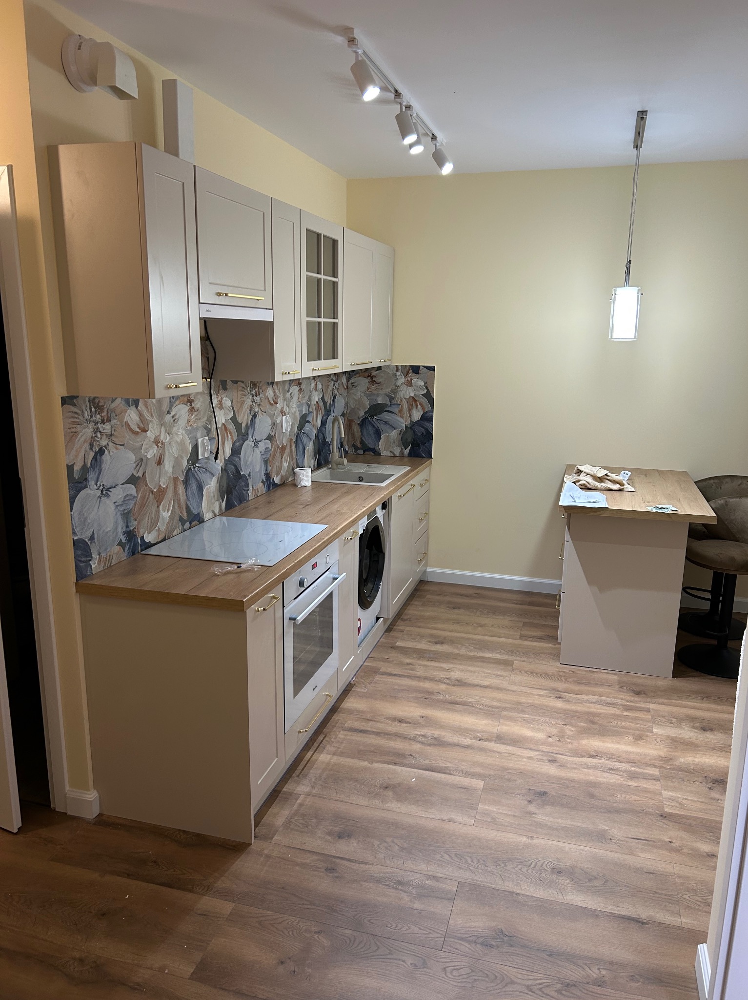 Jasna kuchnia po remoncie: beżowe szafki z drewnianym blatem, kwiatowy panel nad blatem, drewniana podłoga, oświetlenie punktowe i wisząca lampa.