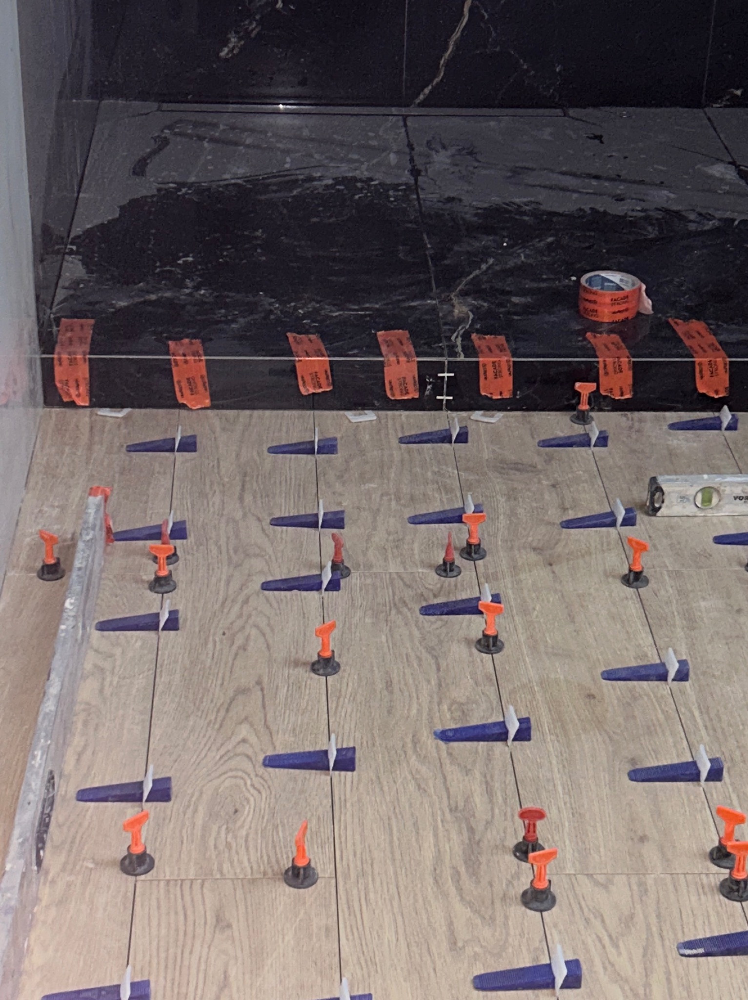 Układanie płytek imitujących drewno z użyciem systemu poziomowania. W tle czarne płytki marmurowe na ścianie. Widoczna poziomica i taśma malarska.