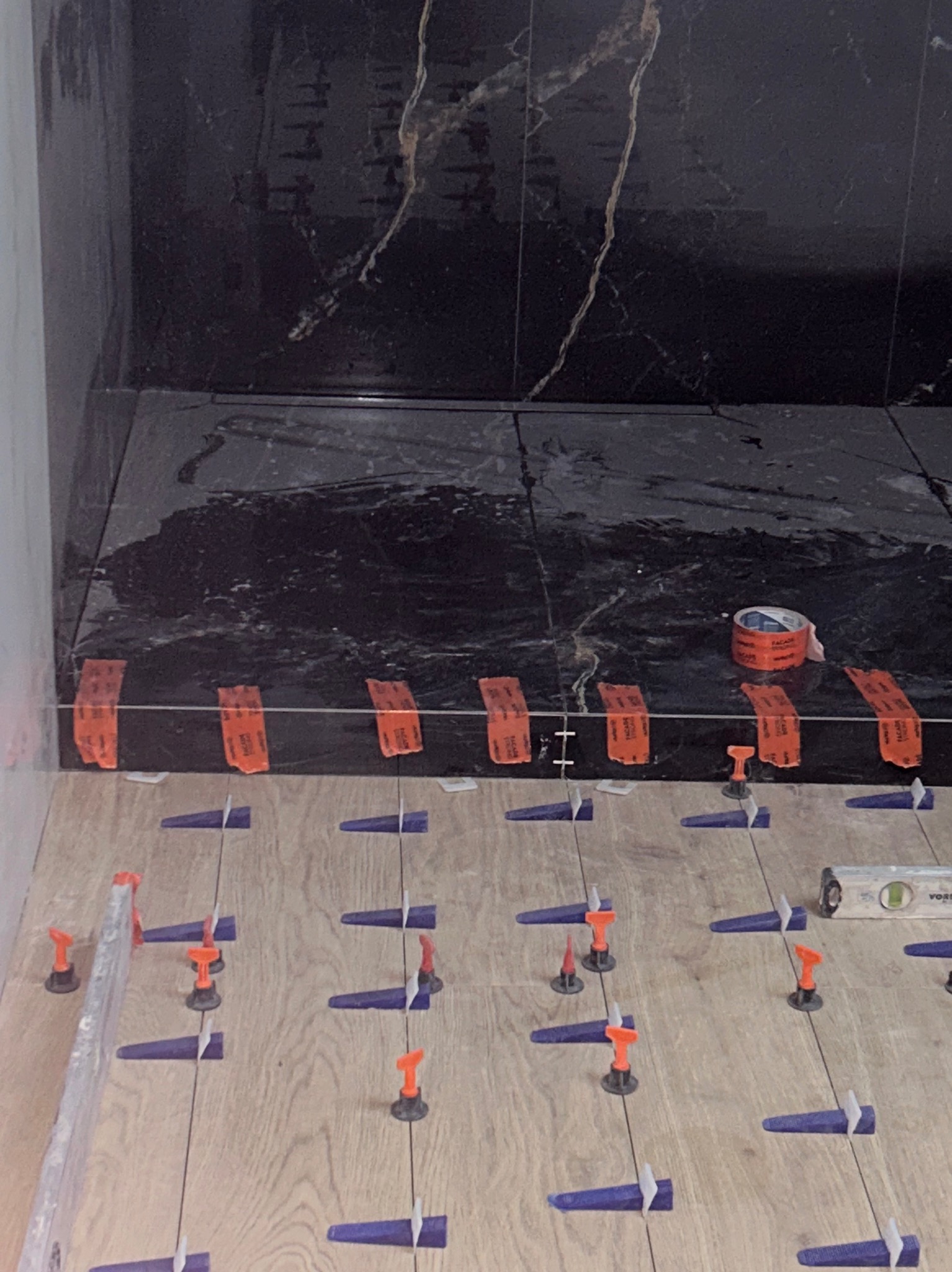 Układanie płytek podłogowych imitujących drewno z użyciem systemu poziomowania. W tle czarne płytki ścienne z marmurkowym wzorem i rolka taśmy.