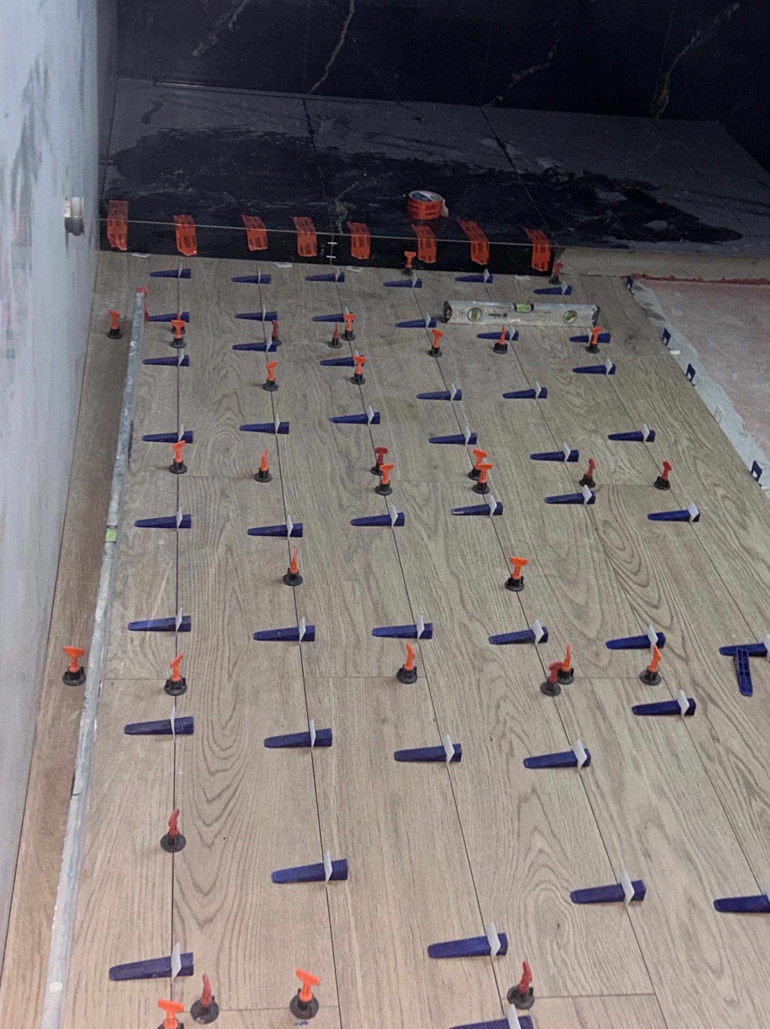 Układanie płytek imitujących drewno z użyciem systemu poziomowania. Widoczny poziomica i czarne płytki na ścianie w tle.