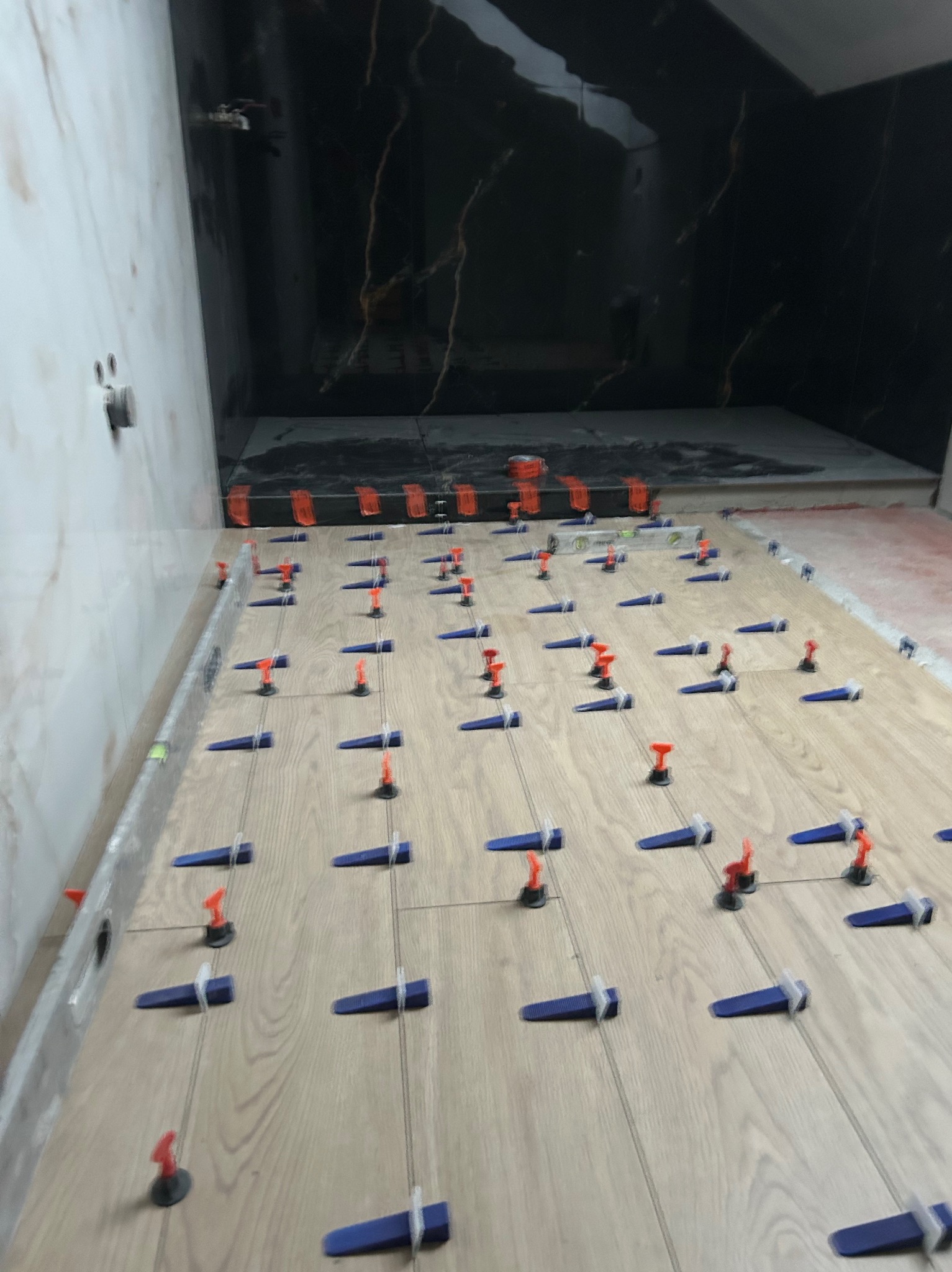 Układanie paneli podłogowych w łazience z użyciem klipsów poziomujących. Widoczny poziomica i fragment ściany z marmurowymi płytkami.