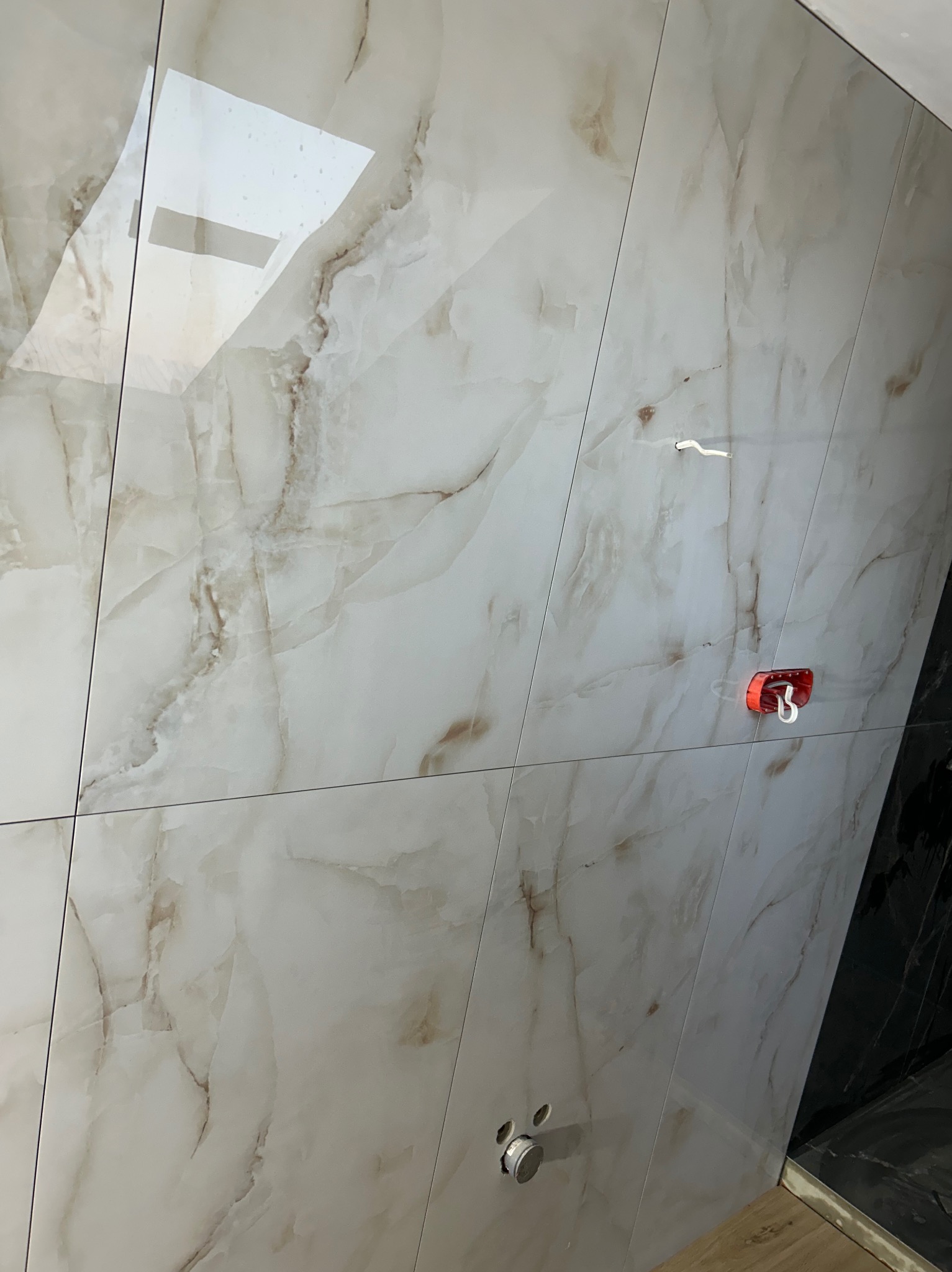 Ściana wyłożona błyszczącymi, beżowo-białymi płytkami imitującymi marmur. Widoczne otwory na armaturę i uchwyt na słuchawkę prysznicową. Fragment czarnej ściany w tle.