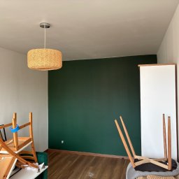 A&P Fach-Remont - Pokój po malowaniu: jedna ściana w odcieniu butelkowej zieleni, reszta biała. Na podłodze panele, w tle lampa z abażurem z naturalnego materiału i meble przygotowane do transportu.