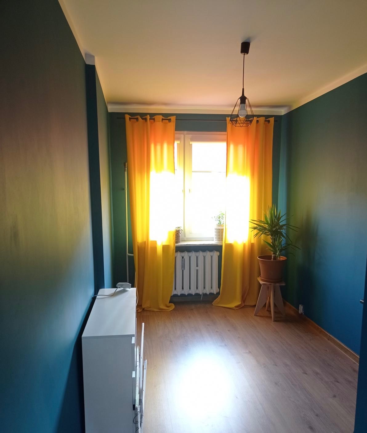 Pokój po odświeżeniu: turkusowe ściany, drewniana podłoga, żółte zasłony w oknie, biały grzejnik i komoda. Na stołku doniczka z rośliną. Nowoczesna lampa sufitowa.
