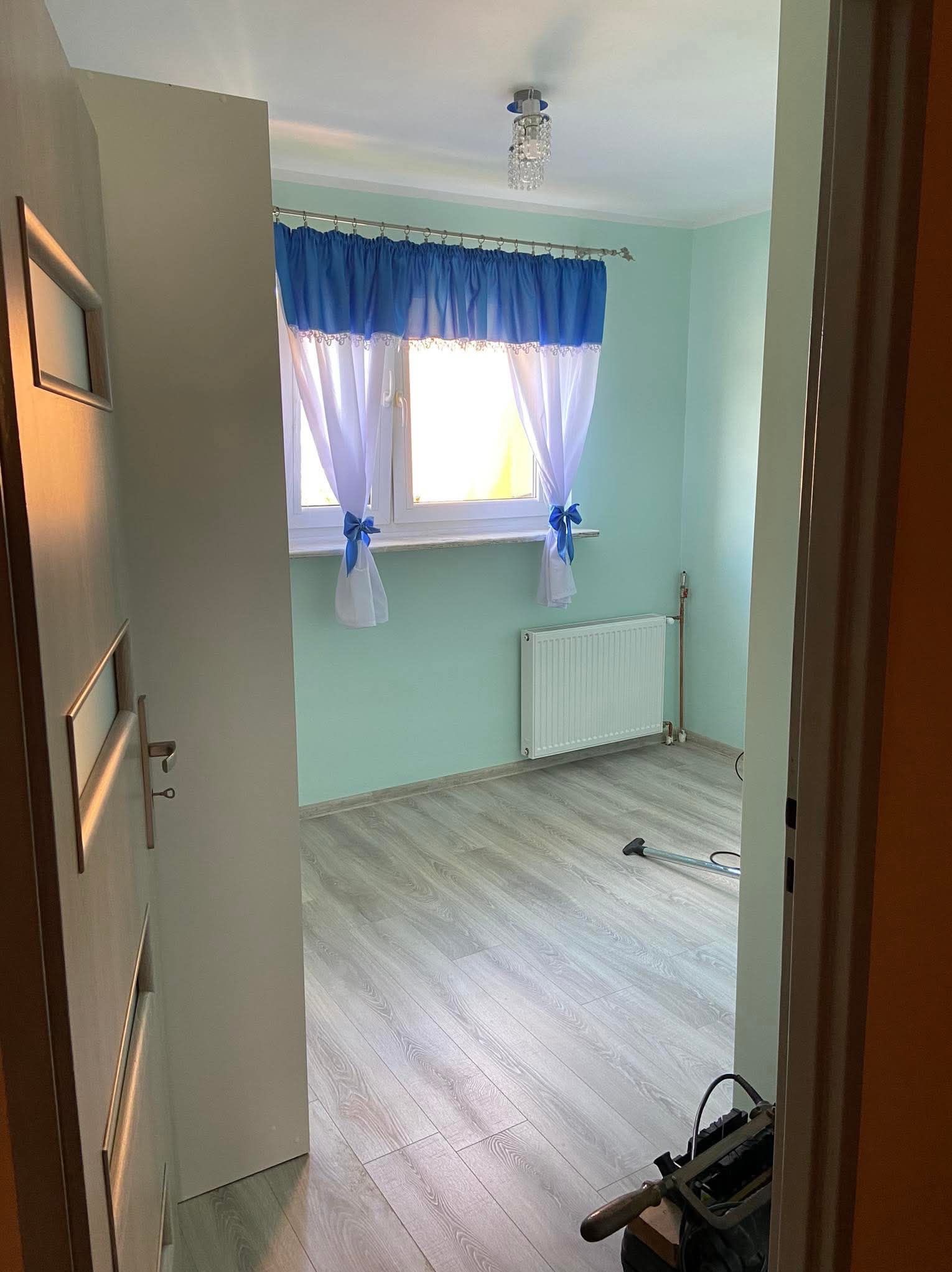 Jasne wnętrze pokoju po odświeżeniu: świeżo pomalowane ściany w odcieniach mięty, panele podłogowe imitujące drewno i okno z niebieskimi zasłonami. Widoczny fragment narzędzi.