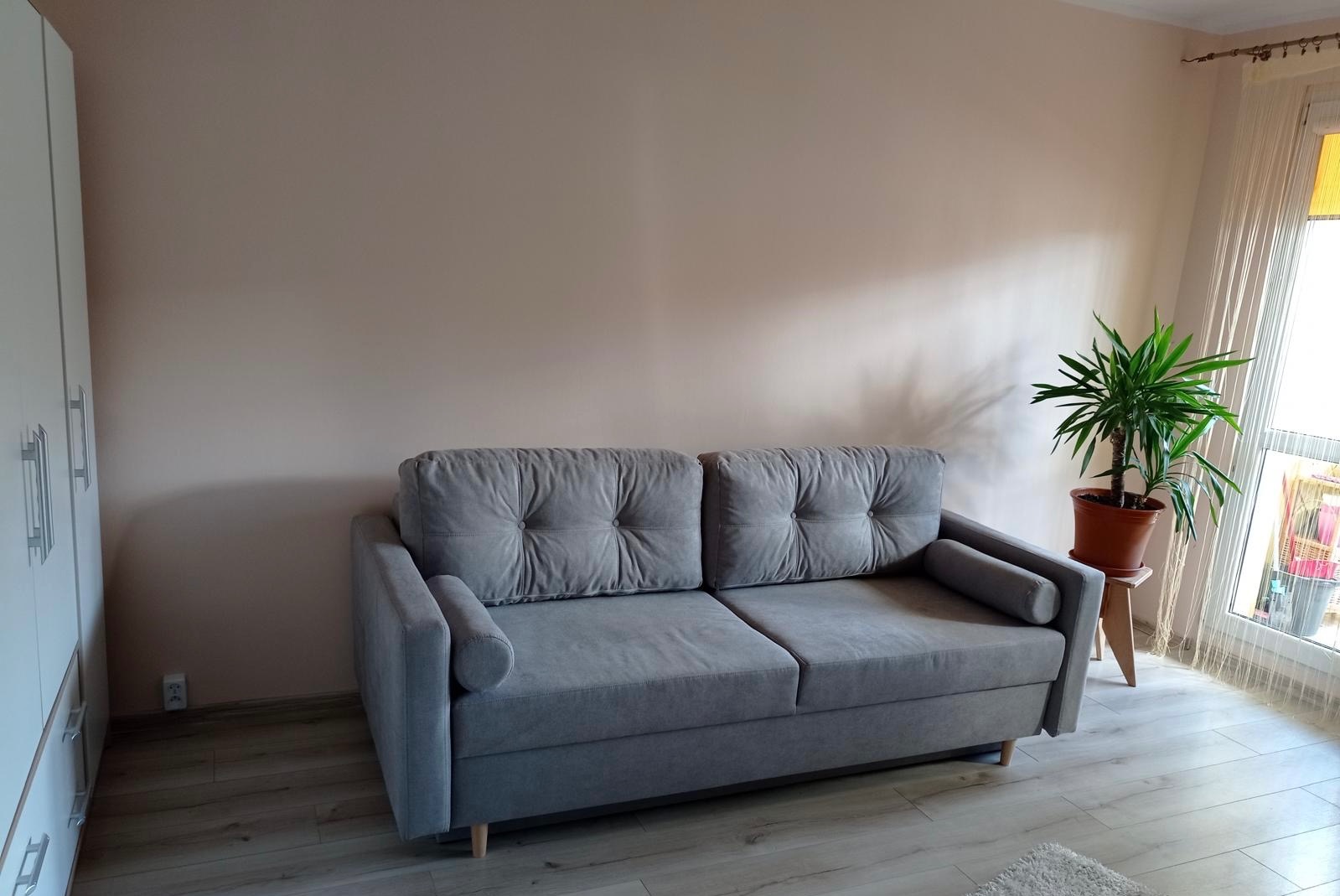 Jasny pokój po odświeżeniu: szara sofa z poduszkami na tle kremowej ściany, obok biała szafa i roślina doniczkowa przy oknie. Podłoga wyłożona panelami.