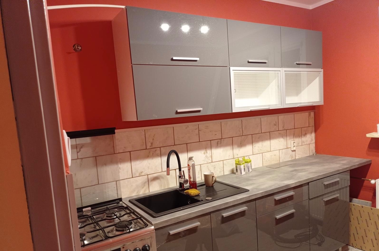 Nowoczesna kuchnia w szarościach i czerwieniach: błyszczące szafki, marmurowe płytki i czarny zlew. Widoczna kuchenka gazowa i akcesoria kuchenne.