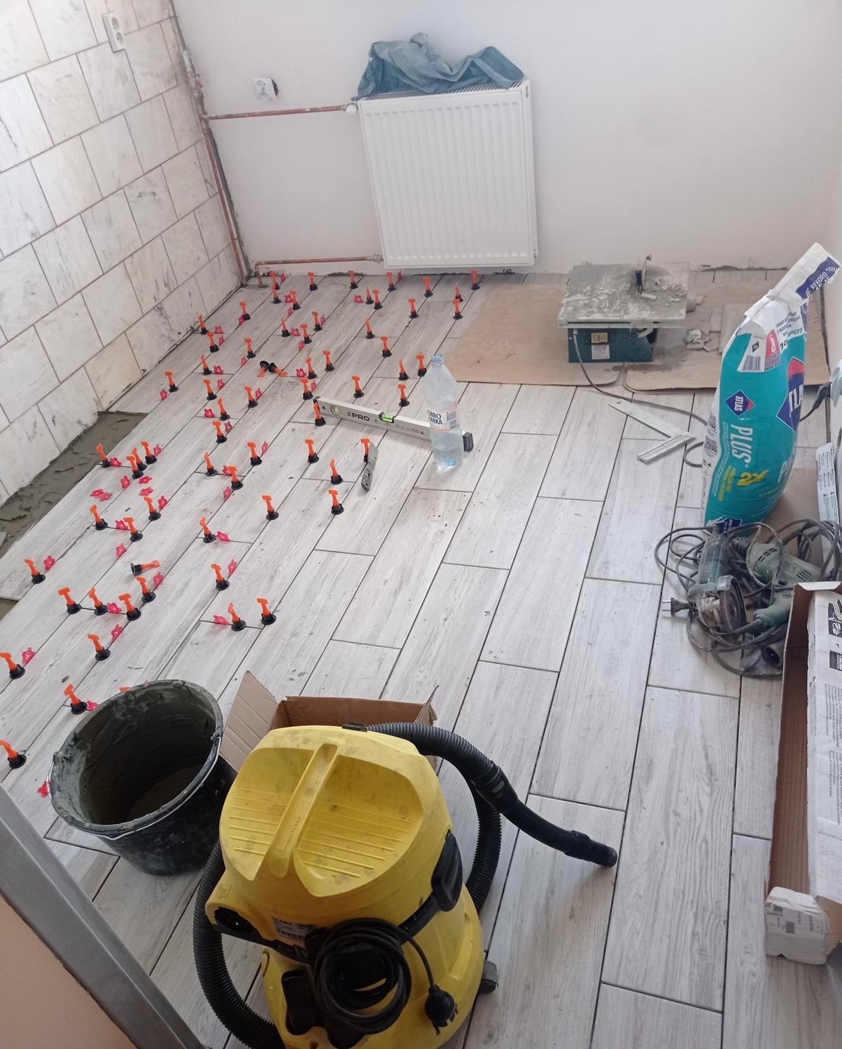 Układanie płytek podłogowych z użyciem systemu poziomowania. Widoczny fragment ściany z ułożonymi płytkami, narzędzia i materiały budowlane. Jasne płytki imitujące drewno.