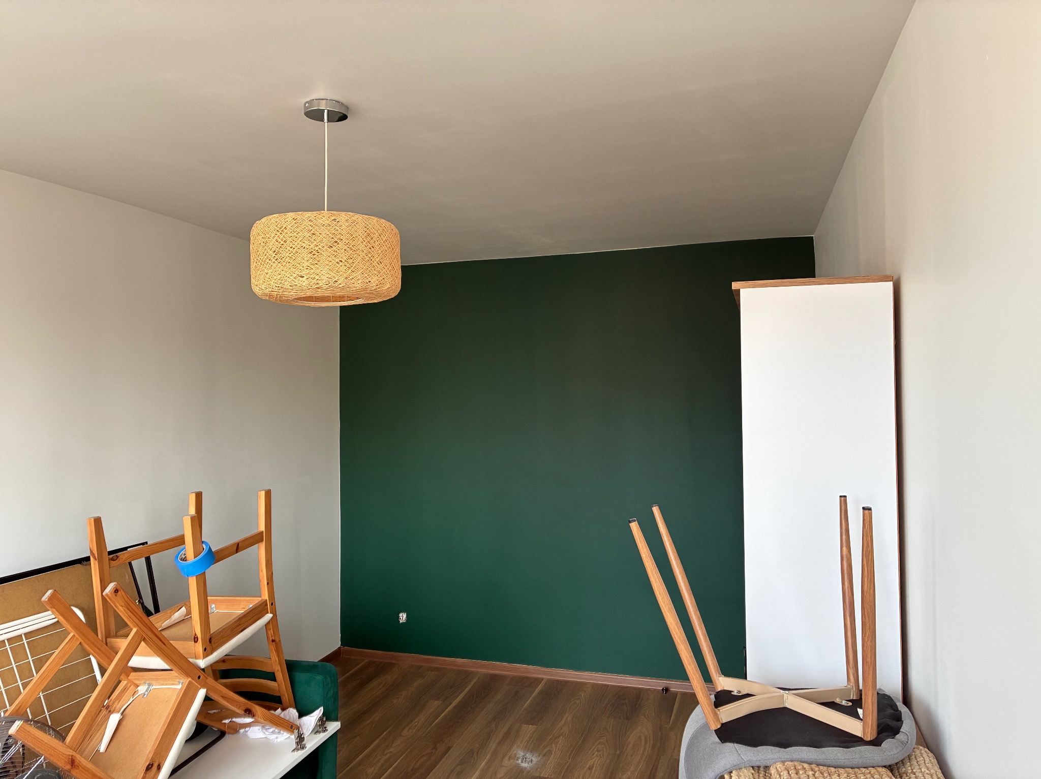 Pokój po malowaniu: jedna ściana w odcieniu butelkowej zieleni, reszta biała. Na podłodze panele, w tle lampa z abażurem z naturalnego materiału i meble przygotowane do transportu.