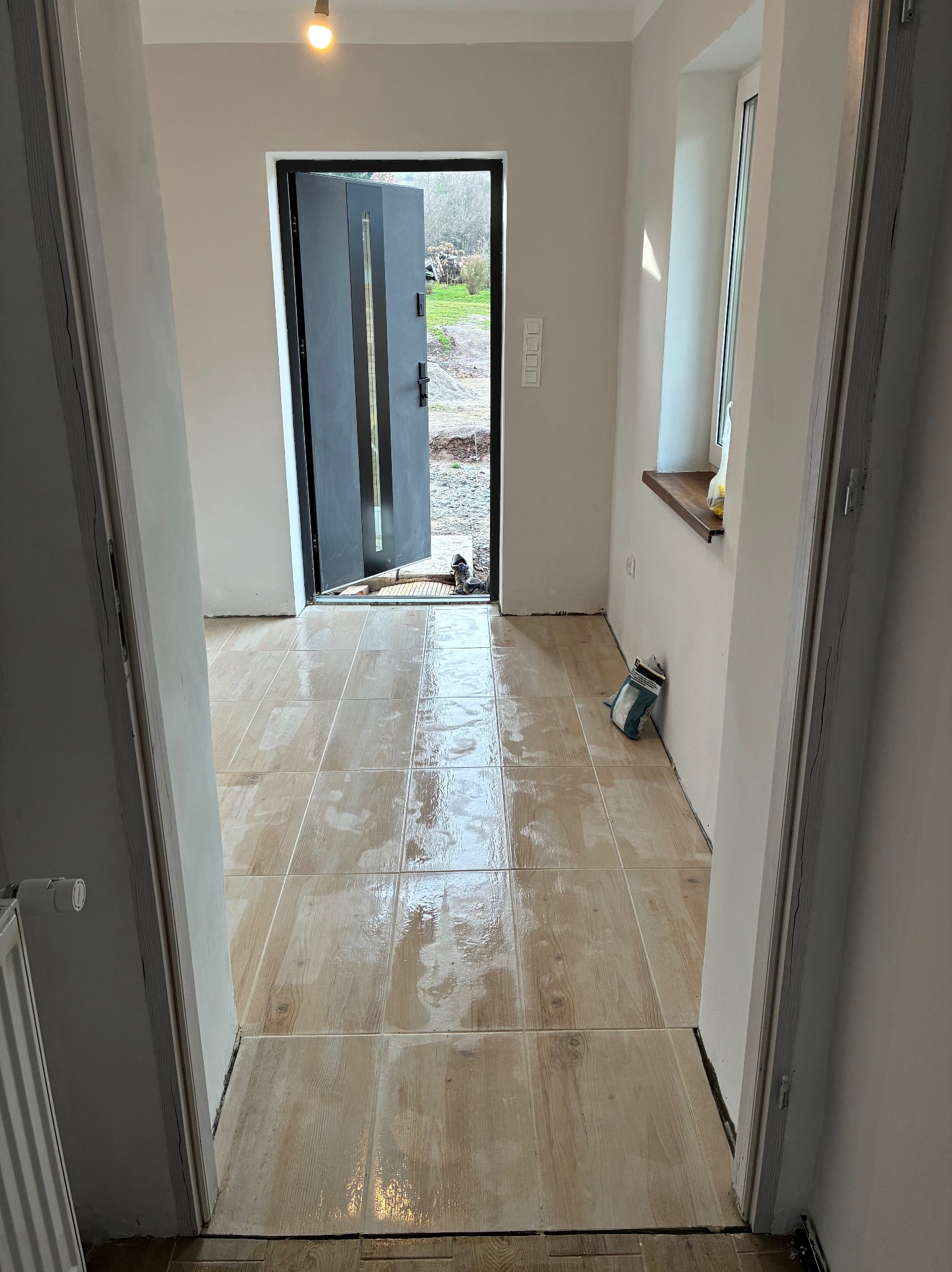 Jasne, drewnopodobne płytki podłogowe w nowym wnętrzu, widok z progu. Otwarta, grafitowa rama drzwi wejściowych, ściany w odcieniach bieli, okno z drewnianym parapetem.