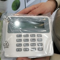 HTControl Hryhorii Tokarenko - Panel sterowania alarmem Safer w folii, trzymany w dłoniach. Biała obudowa z szarymi przyciskami numerycznymi i wyświetlaczem. Widoczne oznaczenia recyklingu na opakowaniu.