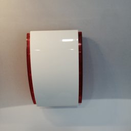 HTControl Hryhorii Tokarenko - Biała obudowa sygnalizatora alarmowego z czerwonymi elementami, zamontowana na jasnej ścianie. Widoczne przewody u góry.