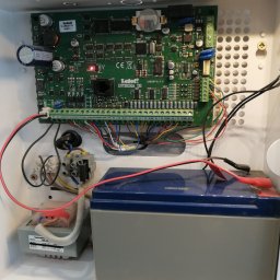 HTControl Hryhorii Tokarenko - Centrala alarmowa Satel Integra 32 w białej obudowie, widoczne podłączenia kabli, akumulator i transformator. Elementy elektroniczne na zielonej płytce drukowanej.