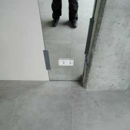 HTControl Hryhorii Tokarenko - Białe podwójne gniazdko elektryczne zamontowane na szarej ścianie, widoczne nogi osoby w odbiciu lustra. Surowy betonowy filar obok.