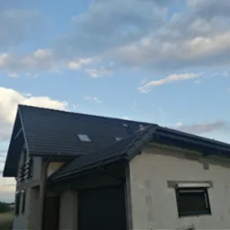 Dom w stanie surowym zamkniętym z ciemną dachówką i oknami. Widoczne orynnowanie i instalacja odgromowa. Błękitne niebo z chmurami w tle.