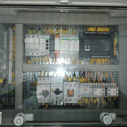 HTControl Hryhorii Tokarenko - Rozdzielnica elektryczna w przezroczystej obudowie, widoczne liczne kable i moduły, oświetlenie odbija się od obudowy. Elementy sterujące i zabezpieczające wewnątrz.