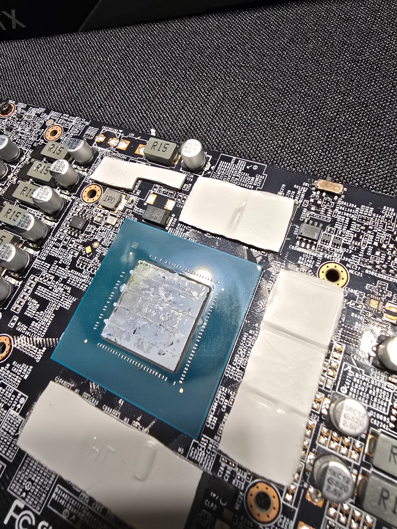 Detale płyty głównej komputera z procesorem i elementami chłodzącymi, widoczne kondensatory i układy scalone na tle szarej tkaniny o drobnej fakturze.
