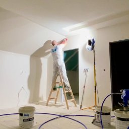 ILYA VLASOV - Malowanie natryskowe wnętrza: mężczyzna na drabinie, białe ściany, sprzęt malarski, wiadro farby Knauf Roll & Spray. Widoczne oświetlenie i przygotowane podłoże.