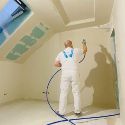 ILYA VLASOV - Pracownik w białym kombinezonie maluje ściany natryskowo w jasnym pokoju na poddaszu z oknem dachowym. Widoczne ślady po szpachlowaniu na suficie i ścianach.