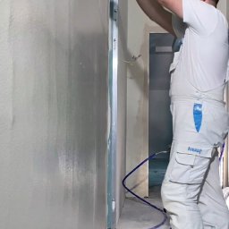 ILYA VLASOV - Pracownik w kombinezonie Knauf wygładza ścianę szpachelką. Widoczne świeżo nałożone warstwy gładzi i niebieski przewód. W tle fragment korytarza w trakcie prac wykończeniowych.