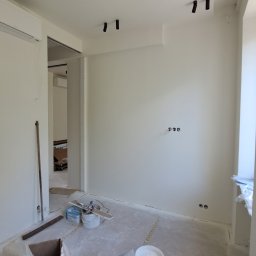 ILYA VLASOV - Wnętrze po malowaniu: białe ściany, czarne reflektory sufitowe, widoczne puszki elektryczne. Na podłodze resztki materiałów budowlanych i narzędzia malarskie.