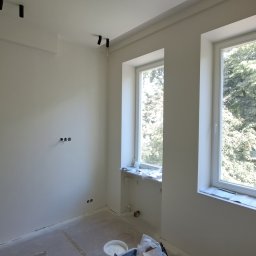 ILYA VLASOV - Jasne wnętrze po malowaniu ścian na biało, widoczne dwa okna z widokiem na zieleń, gładkie ściany z gniazdkami i lampy sufitowe. Na podłodze widoczne materiały malarskie.