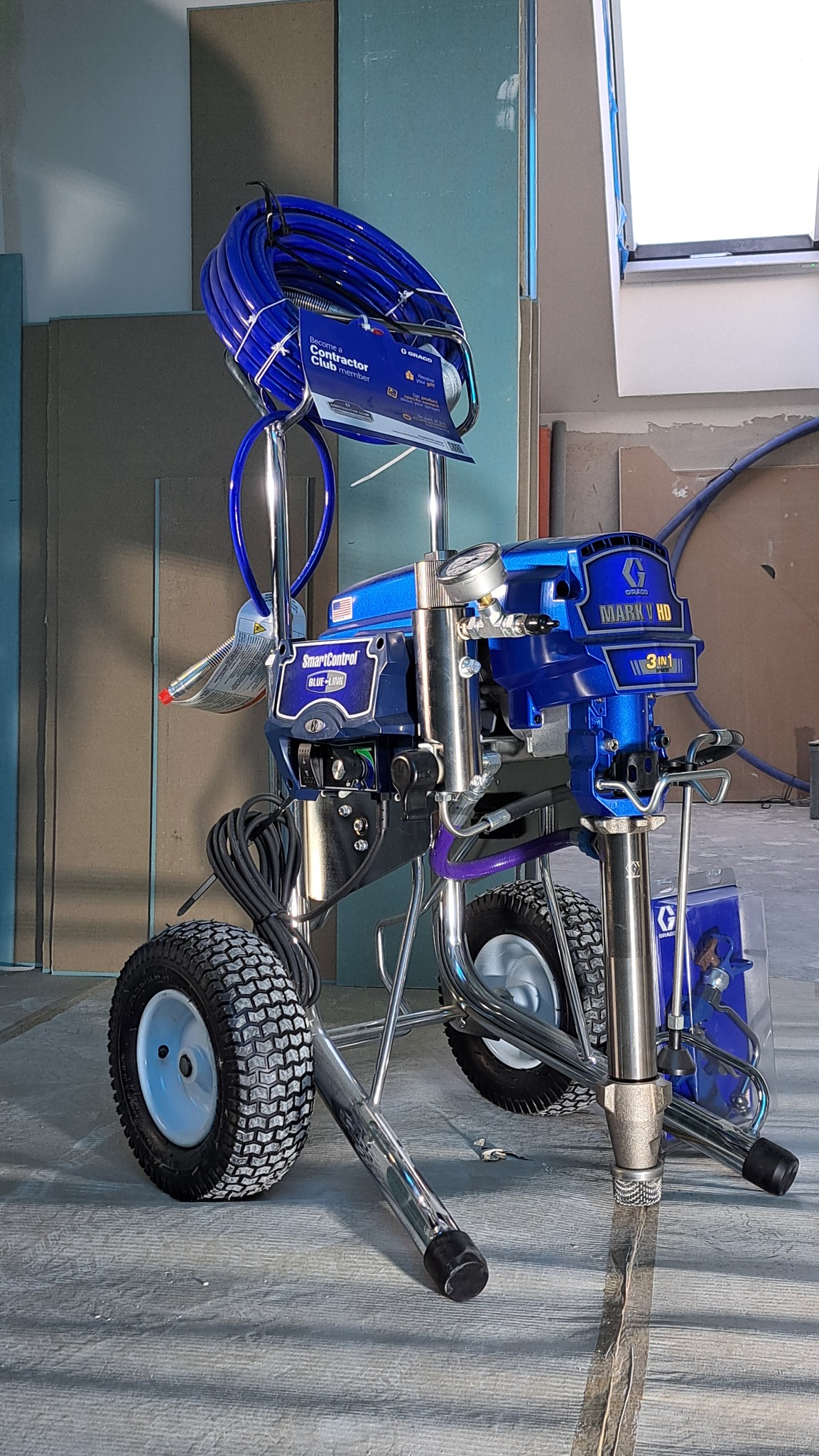 Profesjonalny agregat malarski Graco Mark V HD 3in1 w kolorze niebieskim, z widocznym wężem i elementami sterującymi, na tle surowych ścian z płyt kartonowo-gipsowych.