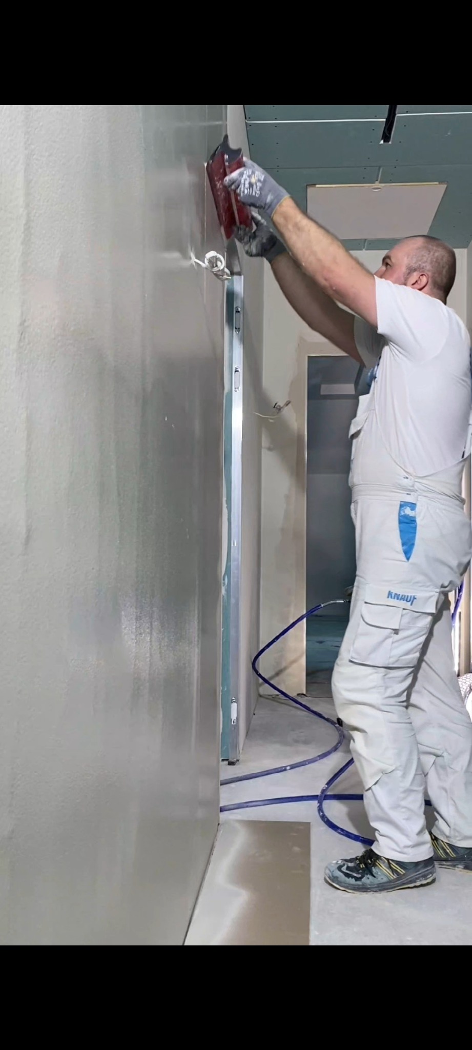 Pracownik w kombinezonie Knauf wygładza ścianę szpachelką. Widoczne świeżo nałożone warstwy gładzi i niebieski przewód. W tle fragment korytarza w trakcie prac wykończeniowych.