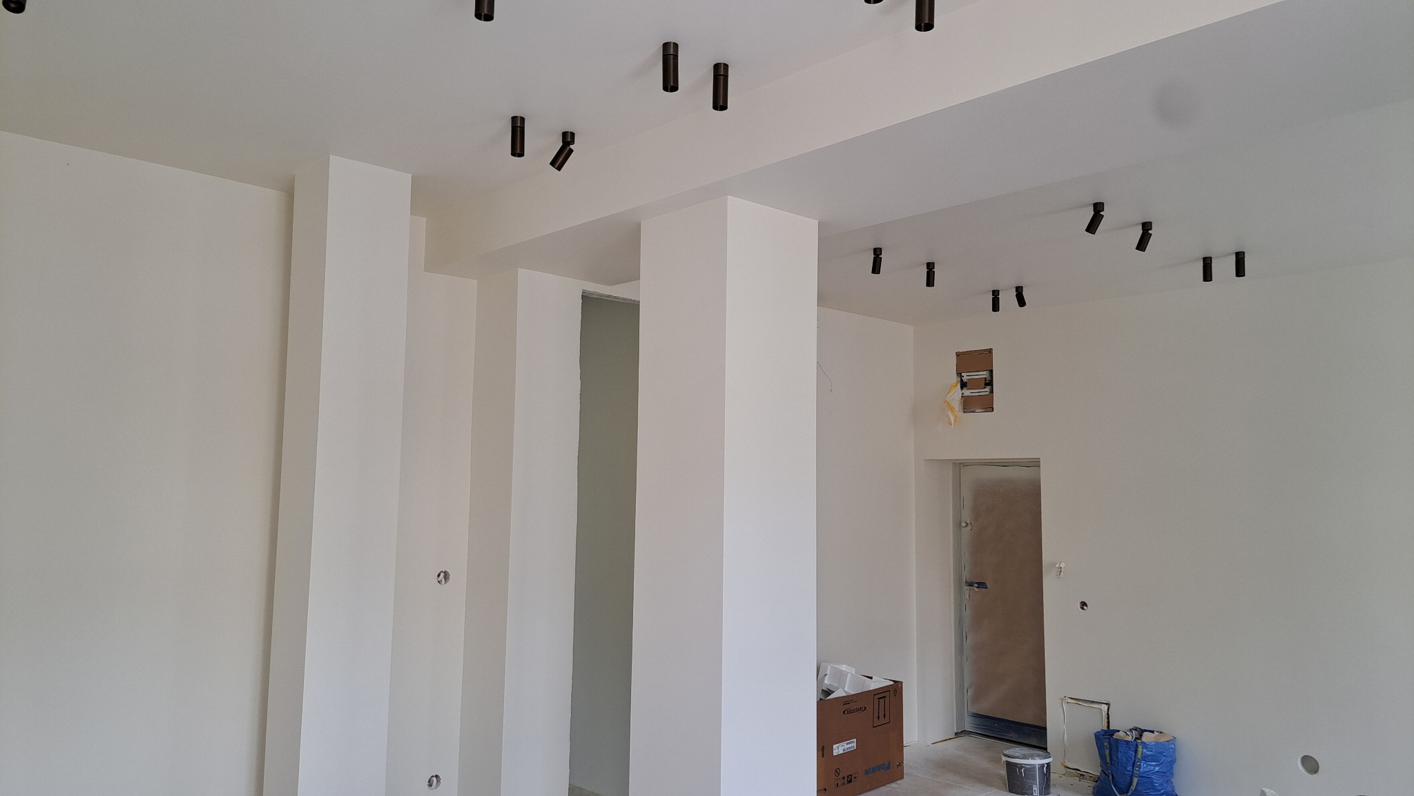Jasne wnętrze po remoncie z białymi ścianami, kolumnami i czarnymi reflektorami sufitowymi. Widoczne ślady po instalacjach elektrycznych i zabezpieczone drzwi.