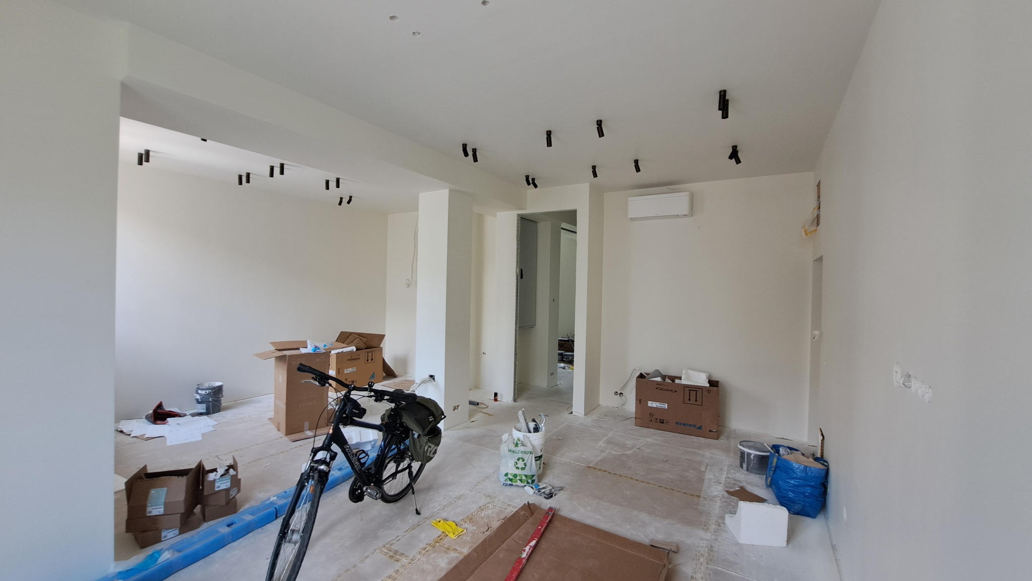 Wnętrze w trakcie prac wykończeniowych: białe ściany, sufit z zamontowanymi punktami oświetleniowymi, kartony, rower i narzędzia malarskie na podłodze.