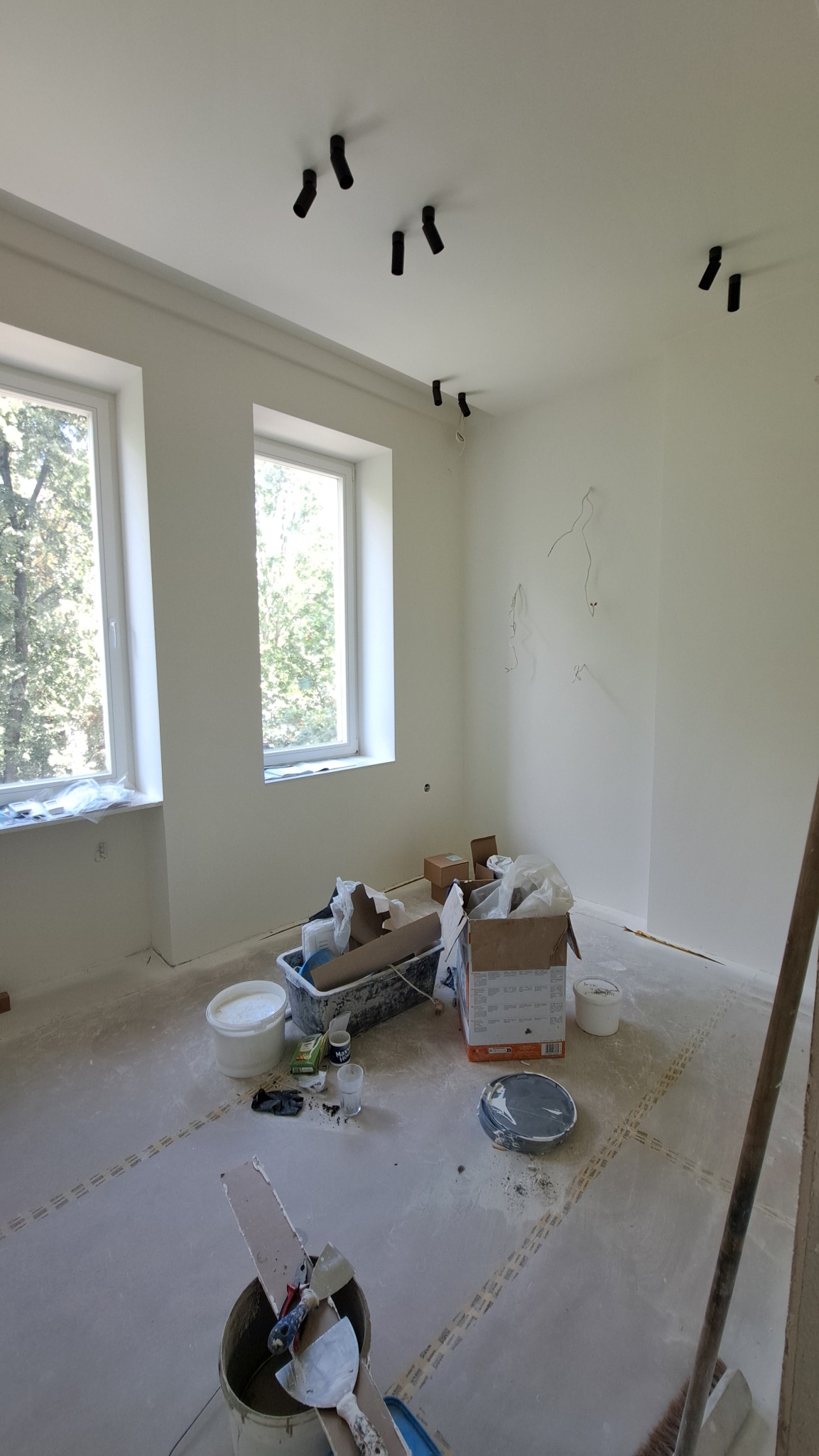 Pokój po gładziowaniu i malowaniu na biało, widoczne dwa okna z widokiem na zieleń, czarne reflektory na suficie i narzędzia malarskie na podłodze.