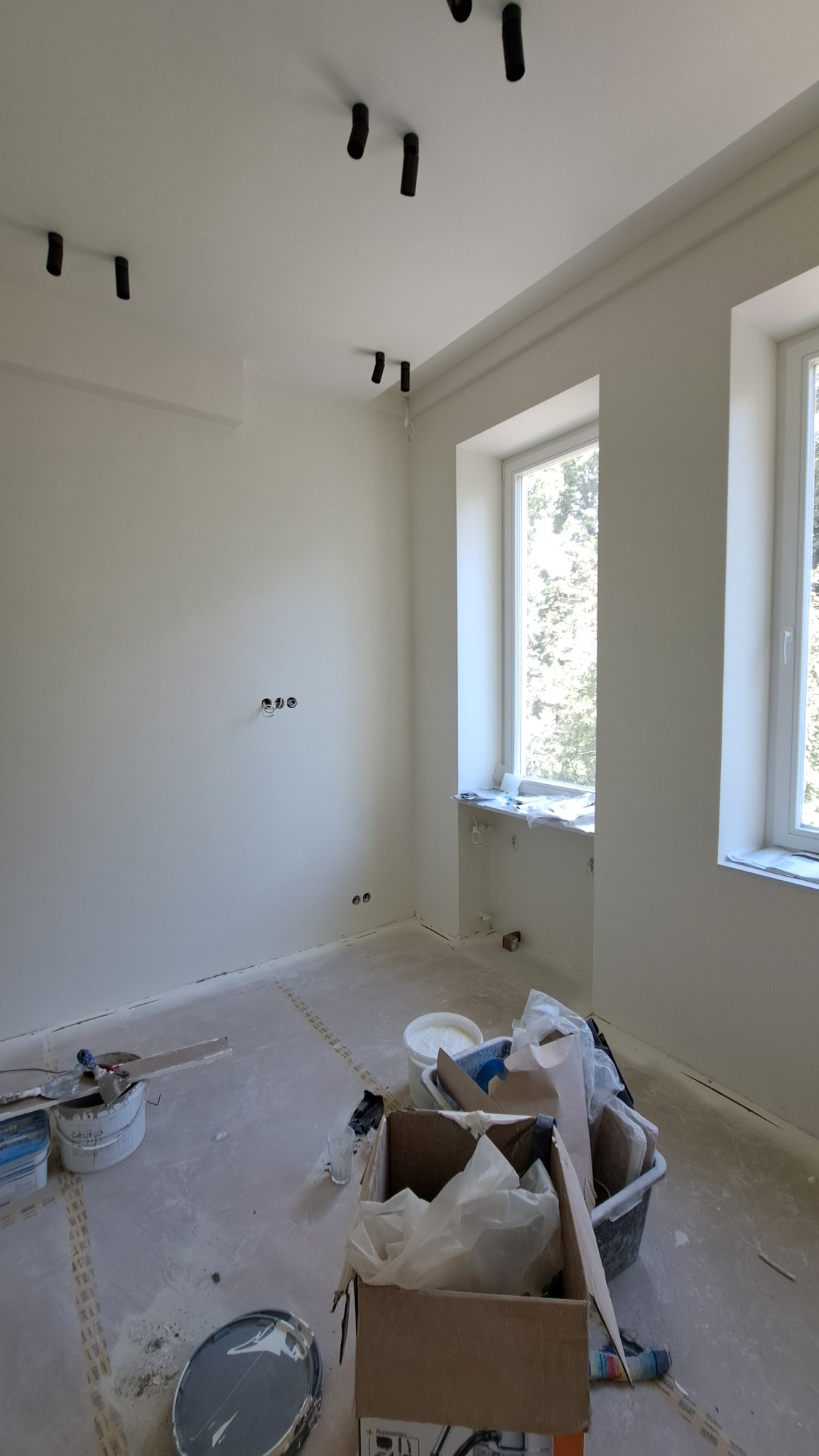 Jasne wnętrze po malowaniu, widoczne puszki z farbą i narzędzia malarskie na podłodze. Świeżo pomalowane ściany i sufit z zamontowanym oświetleniem punktowym.