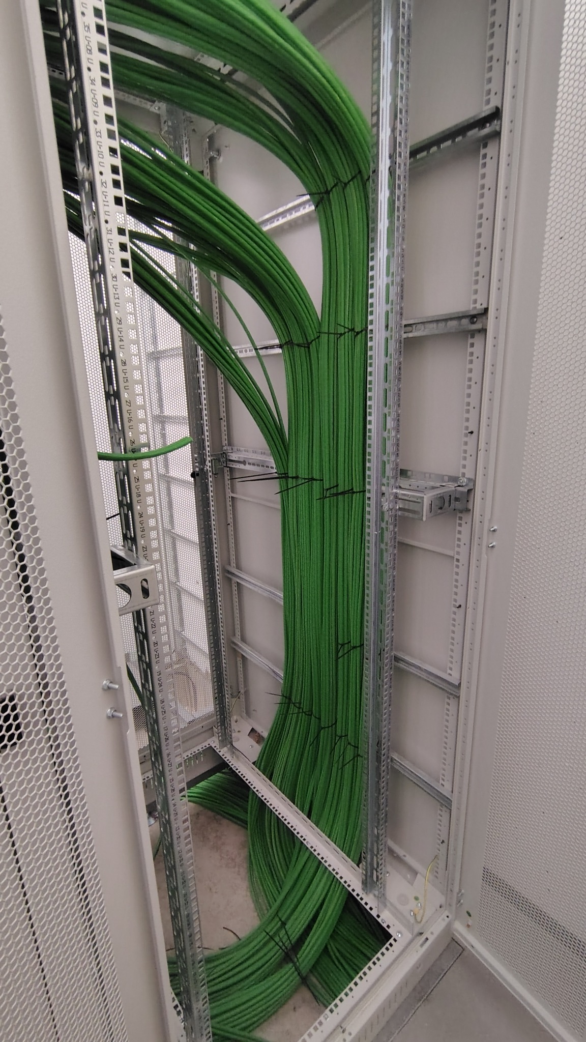 Szafa rack z uporządkowanymi wiązkami zielonych kabli, spiętych czarnymi opaskami. Widoczne metalowe elementy konstrukcyjne szafy.