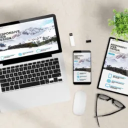 Responsywna strona WWW na laptopie, smartfonie i tablecie, z motywem g&oacute;rskim. Okulary, myszka i roślina doniczkowa na biurku. Prezentacja projektu strony.