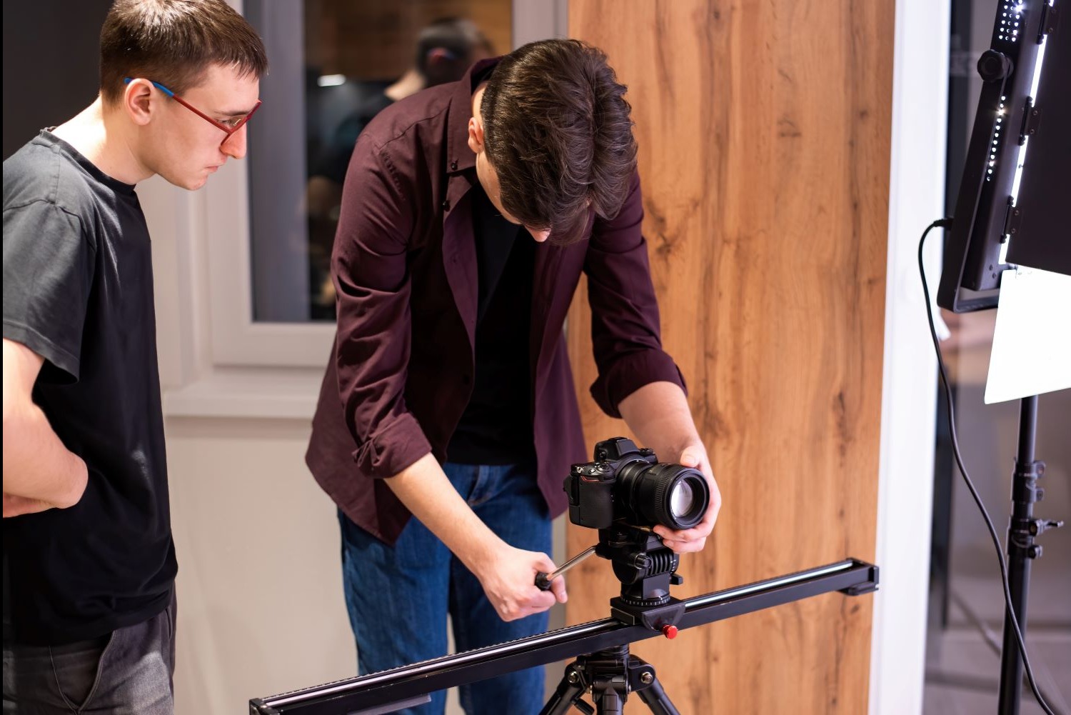 Ekipa filmowa ustawia kamerę na sliderze przed drewnianą ścianą. Jeden z mężczyzn obserwuje, a drugi reguluje ustawienia. W tle lampa oświetleniowa.