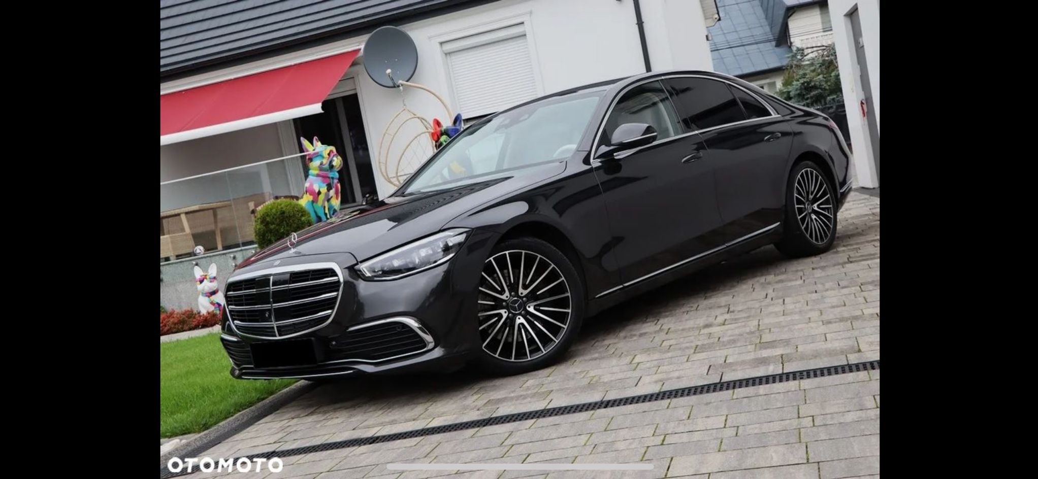 Elegancki, czarny Mercedes S-klasa zaparkowany na posesji z kolorowymi dekoracjami ogrodowymi. Widoczne detale: felgi, chromowane listwy, reflektory.