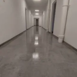 Długi, jasny korytarz z szarymi płytkami podłogowymi i białymi ścianami. Widoczne pionowe rury, drzwi i kwadratowe lampy sufitowe.