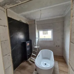 Wnętrze łazienki w trakcie prac: biała wanna wolnostojąca, czarne płytki imitujące marmur, drabina, okno. Widoczne niedokończone ściany z bloczk&oacute;w.