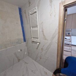SZBF - Łazienka w trakcie wykończenia: marmurowe płytki, biały grzejnik drabinkowy, futryna drzwi. W tle drabina i częściowo widoczna osoba w niebieskiej bluzie.