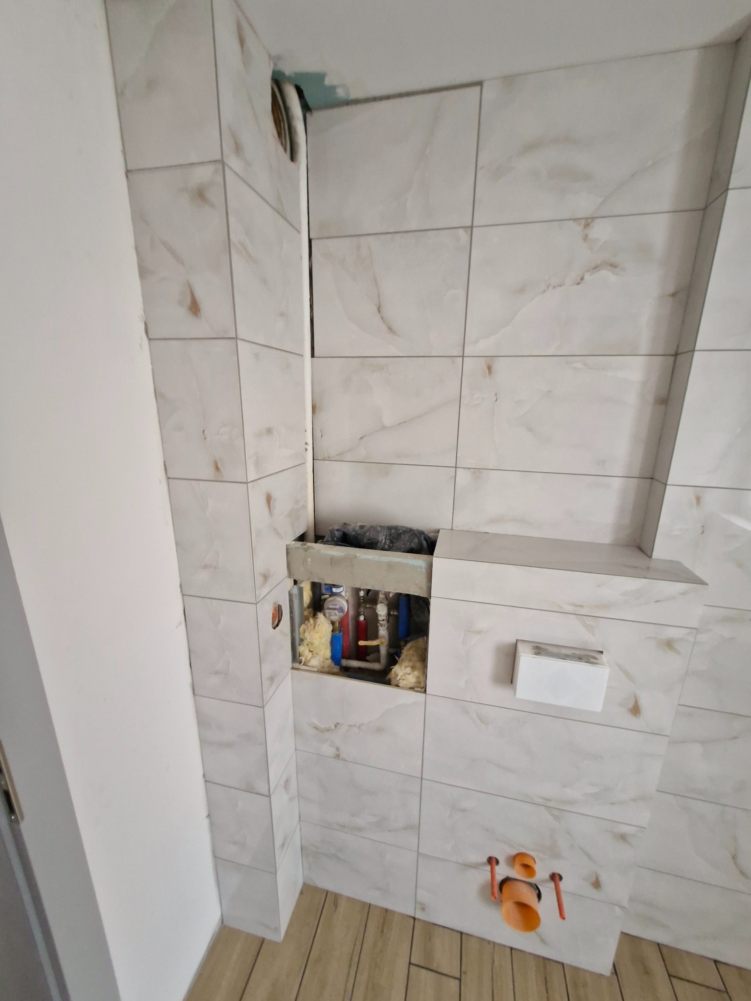 Biała łazienka w trakcie remontu: ułożone płytki imitujące marmur, widoczne rury i instalacja podtynkowa. Częściowo zamontowana spłuczka WC.